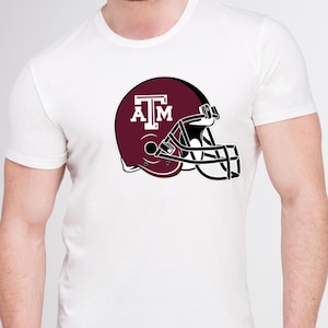 May include: White t-shirt featuring a maroon football helmet graphic with the letters "A" and "M" in white. The helmet has a black face mask. The shirt is a classic crew neck style.