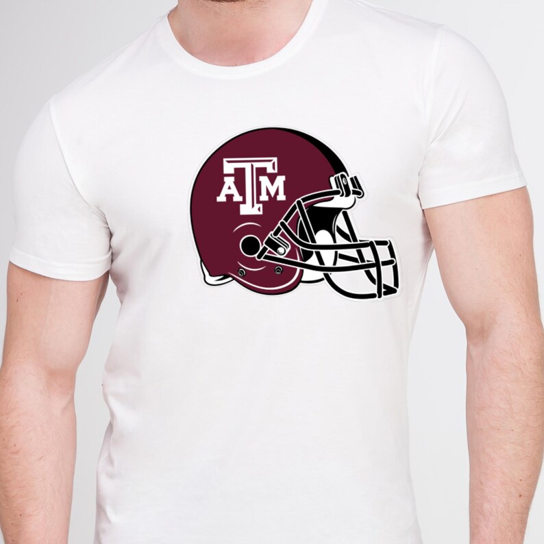 May include: White t-shirt featuring a maroon football helmet graphic with the letters "A" and "M" in white. The helmet has a black face mask. The shirt is a classic crew neck style.