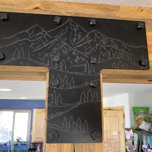 Può includere: Staffa a T in metallo nero con una scena di montagna e foresta incisa sulla superficie. La staffa è fissata a una trave di legno con più viti.