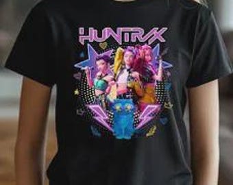May include: Black t-shirt featuring a graphic of three stylized figures with the text "HUNTRAX" above them. The graphic includes stars, lightning bolts, and a blue cat-like creature. The figures are in vibrant colors.
