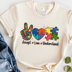 May include: A cream-colored t-shirt with a peace sign made of puzzle pieces, a heart made of puzzle pieces, and the text "Accept * Love * Understand".