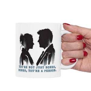May include: White ceramic mug with a black silhouette of a couple looking at each other. The text "You're not just bones, bones, you're a person." is printed in blue on the mug.