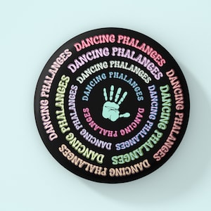 Puede incluir: Un botón redondo negro con las palabras "DANCING PHALANGES" en un arcoíris de colores. El centro presenta una huella de mano azul claro.