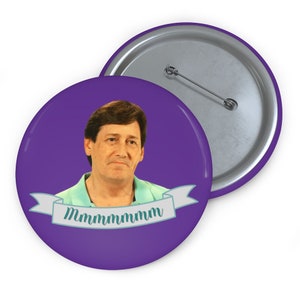 May include: A purple button with a picture of a man with brown hair and a light blue shirt. The man is looking at the camera with a neutral expression. The button has a white banner with the text "Mmmmmm" in teal.