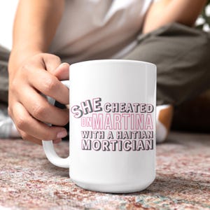 White ceramic mug with bold pink and black text reading “She cheated on Martina with a Haitian mortician,” referencing a Real Housewives of Miami quote.