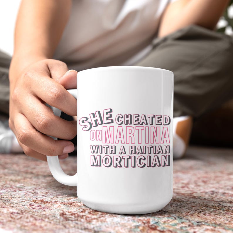 White ceramic mug with bold pink and black text reading “She cheated on Martina with a Haitian mortician,” referencing a Real Housewives of Miami quote.