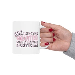 White ceramic mug with bold pink and black text reading “She cheated on Martina with a Haitian mortician,” referencing a Real Housewives of Miami quote.