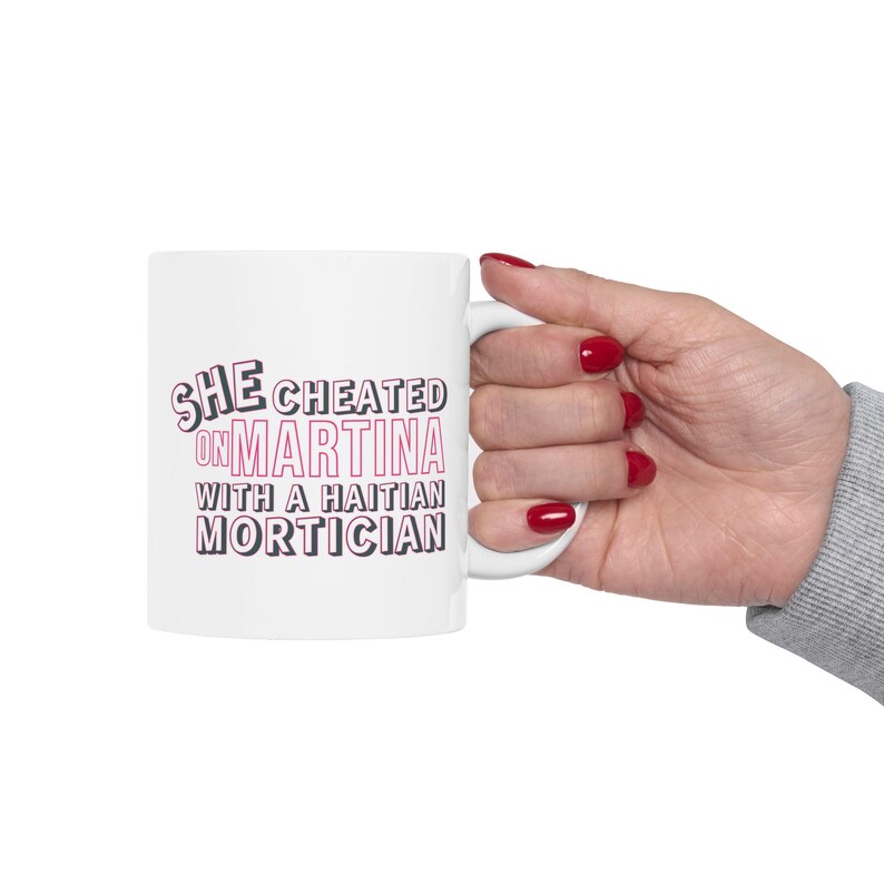 White ceramic mug with bold pink and black text reading “She cheated on Martina with a Haitian mortician,” referencing a Real Housewives of Miami quote.