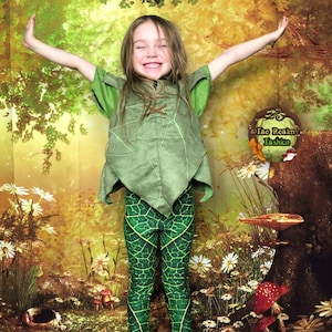 May include: A young person wearing a green leaf-patterned costume with a brown leather belt and matching green leaf-patterned leggings. The costume is reminiscent of a woodland creature. The person is standing on a wooden platform in front of a backdrop of a forest scene with mushrooms and flowers.