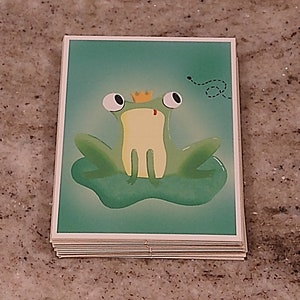 May include: A green frog wearing a gold crown sits on a lily pad with a small fly buzzing around it. The frog has large, white eyes and a red mouth. The background is a light green.