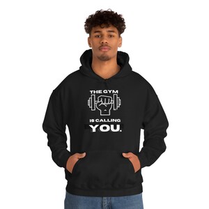 Könnte beinhalten: Schwarzer Kapuzenpullover mit einem weißen Aufdruck, der "The Gym is Calling You." lautet. Die Grafik enthält ein stilisiertes Bild einer Hantel und einer Faust.