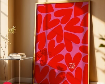 Könnte beinhalten: Ein gerahmter Kunstdruck mit einem Muster aus roten Herzen auf rosa Hintergrund. Die Worte "LOVE LIVES HERE" sind weiß gedruckt. Der Rahmen ist goldfarben.