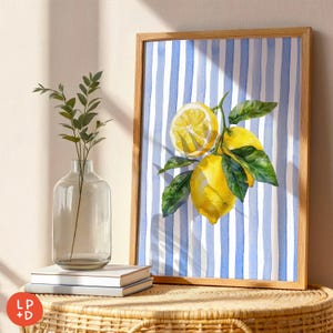May include: A framed watercolor print featuring lemons and green leaves against a blue and white striped background. The artwork is in a light brown wooden frame. A clear glass vase with greenery and a stack of books are on a woven table.