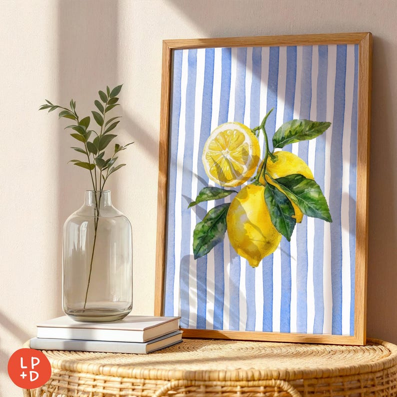 May include: A framed watercolor print featuring lemons and green leaves against a blue and white striped background. The artwork is in a light brown wooden frame. A clear glass vase with greenery and a stack of books are on a woven table.