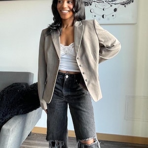 May include: A woman wearing a light brown blazer over a white lace cami top and black jeans with distressed knees.