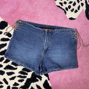 Op de afbeelding: Blauwe spijkerbroekshorts met bruine veters aan de zijkanten. De shorts hebben een knoopsluiting en een klassiek ontwerp met vijf zakken. De denim heeft een medium wassing. Getoond op een roze en zwart patroon oppervlak.