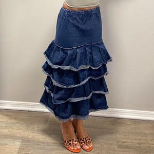 Könnte beinhalten: Dunkelblaue Jeansrock mit mehreren Rüschen. Der Rock hat eine hohe Taille und reicht bis zu den Knöcheln. Das Model trägt Sandalen mit Leopardenmuster und orangefarbenen Sohlen. Der Rock ist eine stilvolle Ergänzung für jede Garderobe.