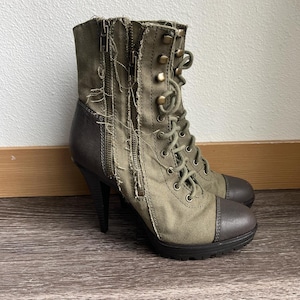 Può includere: Stivaletti alla caviglia in tela verde oliva e pelle marrone con tacco alto. Gli stivali presentano un design stringato, cerniere laterali e un dettaglio in tessuto sfilacciato e sfilacciato. La punta e il tacco sono in pelle marrone.