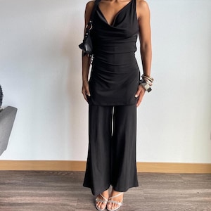May include: A black sleeveless jumpsuit with a cowl neck and wide-leg trousers. The jumpsuit is made of a stretchy fabric and has a fitted bodice. The model is wearing silver sandals.