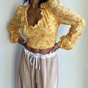 K&ouml;nnte beinhalten: Eine gelbe Bluse mit floralem Muster, langen &Auml;rmeln und einem R&uuml;schenkragen. Die Bluse ist kurz geschnitten und hat eine figurbetonte Silhouette.