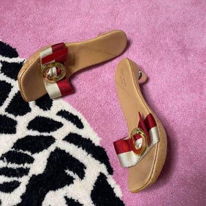 May include: A pair of Kate Spade sandals with a cork sole and a red, white, and gold accent. The sandals have a bow detail with a gold buckle. The shoes are on a pink rug with a black and white pattern.