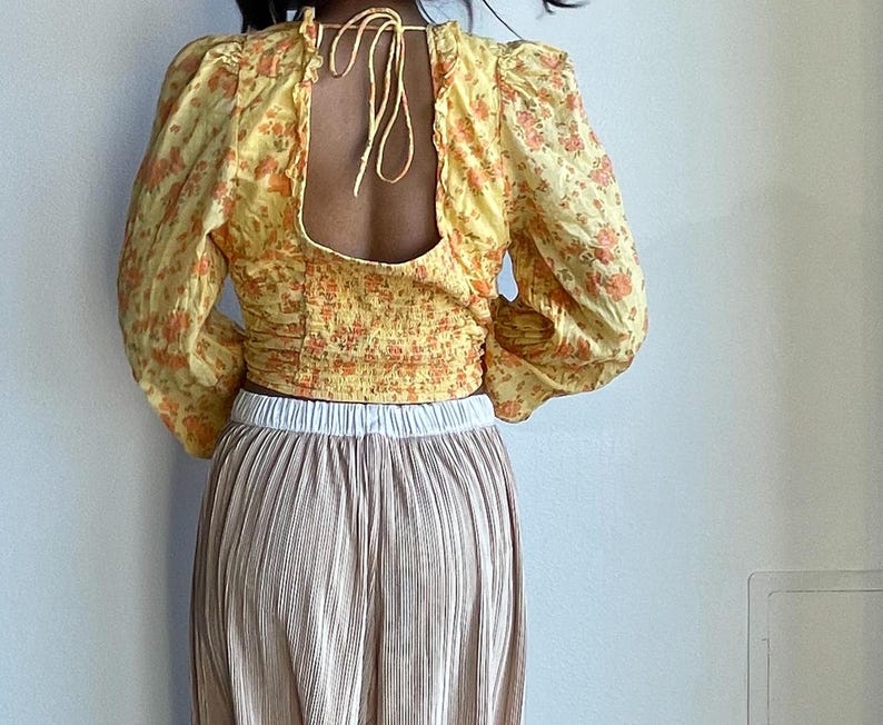 K&ouml;nnte beinhalten: Ein gelbes Oberteil mit Blumendruck, Crop-Top, tiefem R&uuml;cken und langen &Auml;rmeln. Das Oberteil wird mit einem plissierten Rock in hellbrauner Farbe kombiniert.