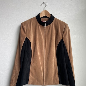 Könnte beinhalten: Hellbraune Jacke mit schwarzen Seiteneinsätzen und einem schwarzen Kragen, hängt an einem Holzkleiderbügel. Die Jacke hat einen durchgehenden Reißverschluss und eine weiche, Wildleder-ähnliche Textur. Neutrale Farbe.