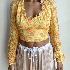 K&ouml;nnte beinhalten: Ein gelbes Crop-Top mit floralem Muster, langen &Auml;rmeln und einem R&uuml;schenkragen. Das Oberteil ist in der Taille gerafft und hat eine lockere Passform.
