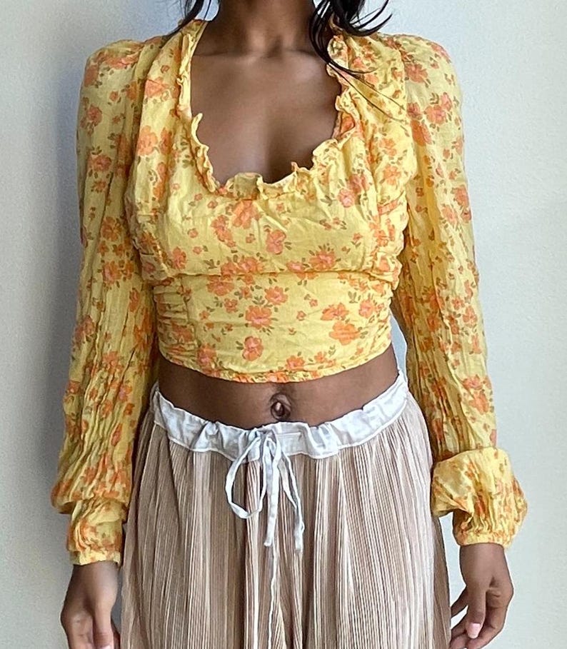 K&ouml;nnte beinhalten: Ein gelbes Crop-Top mit floralem Muster, langen &Auml;rmeln und einem R&uuml;schenkragen. Das Oberteil ist in der Taille gerafft und hat eine lockere Passform.