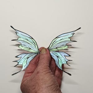 Op de afbeelding: Een paar iriserende elfen vleugels met een lichtblauw en groen kleurenschema. De vleugels worden door een hand tegen een witte achtergrond gehouden. De vleugels hebben zwarte contouren en glitteraccenten.