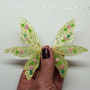 May include: A pair of decorative fairy wings with a glittery, translucent design. The wings are primarily yellow-green with gold glitter accents and feature small pink roses and green leaves. The wings are held against a white background.