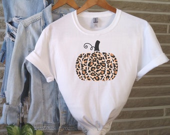 May include: White t-shirt featuring a pumpkin design with a leopard print pattern. The pumpkin has a black stem and a curly vine. The shirt is hanging on a wooden hanger with a denim jacket.