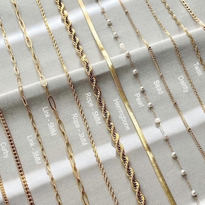 May include: A close-up of ten different gold chains, including a curb chain, a rope chain, a herringbone chain, a link chain, a figaro chain, a twist chain, a dainty chain, a bead chain, a pearl chain, and a vine chain. The chains are laid out on a white surface.