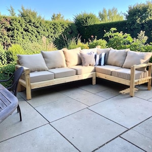 Puede incluir: Un sofá seccional de exterior en forma de L con cojines beige y cojines decorativos. El sofá está sobre un patio de piedra, rodeado de vegetación. Los cojines y almohadas ofrecen comodidad y estilo. Ideal para un jardín.