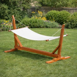 Puede incluir: Un soporte de hamaca de madera con una hamaca blanca estirada entre dos soportes verticales. El soporte es de madera teñida y está sobre un césped. La hamaca está suspendida por una cuerda blanca.