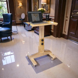 May include: A light-coloured wooden laptop desk on wheels. The desk has a rectangular top with a curved edge, a vertical adjustable support, and a base with four small wheels. A laptop, a black tumbler, and a mouse are on the desk.