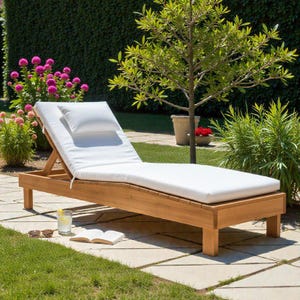 Op de afbeelding: Een houten ligstoel met een wit kussen en kussen. De ligstoel is gemaakt van lichtbruin hout en staat op een stenen terras. Een glas water, een zonnebril en een boek liggen in de buurt. De ligstoel is ongeveer 190 cm lang.