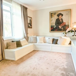 Könnte beinhalten: Eine eingebaute weiße Sitzbank mit mehreren dekorativen Kissen in verschiedenen Farben und Texturen. Die Bank befindet sich in einer Ecke in der Nähe eines Fensters mit beigen Vorhängen. Ein großes Gemälde hängt an der Wand über der Bank.