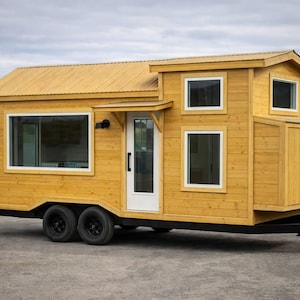 以下が含まれることがあります： 車輪のついた小さな黄色のタイニーハウス。家は切妻屋根、複数の窓、白いドアがあります。トレーラーは黒い車輪と黒いフレームを持っています。外装は木製です。