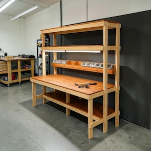 May include: A wooden workbench with a light-colored frame and a darker wood tabletop. The workbench has multiple shelves, including a top shelf and a lower shelf. A light fixture is mounted above the workbench. Tools are visible on the tabletop.