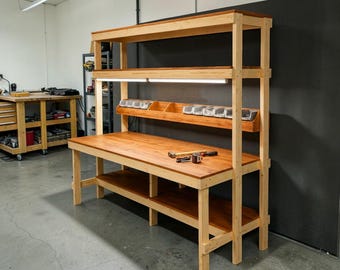 May include: A wooden workbench with a light-colored frame and a darker wood tabletop. The workbench has multiple shelves, including a top shelf and a lower shelf. A light fixture is mounted above the workbench. Tools are visible on the tabletop.