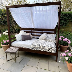 May include: A dark brown wooden outdoor daybed with a canopy, white sheer curtains, and a mattress. Decorative pillows and a floral patterned blanket are arranged on the bed. A small gray upholstered stool sits in front of the bed.