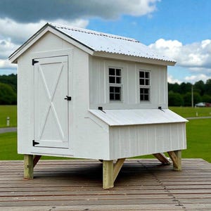May include: A white wooden chicken coop with a corrugated metal roof. The coop has a door, two small windows, and is elevated on wooden posts. The structure is set on a wooden deck, with a green field and blue sky in the background.