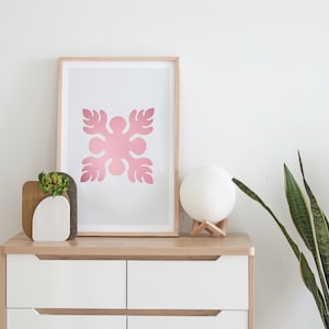 Op de afbeelding: Een ingelijst afdruk van een roze bloemenontwerp in een wit frame op een houten dressoir met een witte keramische lamp en een potplant.