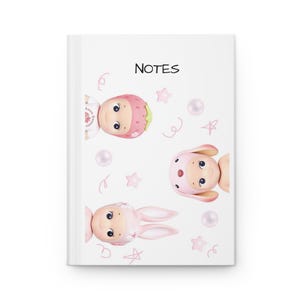 Puede incluir: Cuaderno blanco con la palabra "NOTES" en negro. La portada presenta ilustraciones de tres muñecas de dibujos animados, una con sombrero de fresa y dos con orejas de conejo. Estrellas rosas, perlas y espirales decoran la portada.