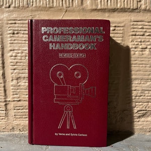 Könnte beinhalten: Ein weinrotes Buch mit dem Titel "PROFESSIONAL CAMERAMAN'S HANDBOOK REVISED EDITION" mit silberner Schrift und einer silbernen Illustration einer alten Filmkamera auf einem Stativ. Das Buch ist von Verne und Sylvia Carlson.