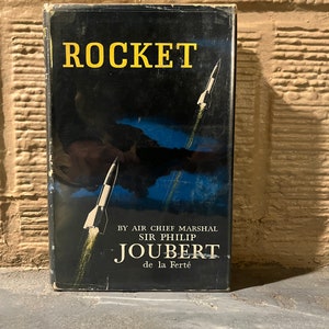 May include: Vintage book cover titled "Rocket" with a black background and yellow lettering. The cover features illustrations of rockets ascending, with the author's name and title below. The book is set against a brick wall.