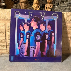 May include: A vintage vinyl record album cover for the band Devo. The cover features a group of men in blue shirts and gray pants. The album title "DEVO" is in large white letters at the top. The album is displayed on a stone surface.