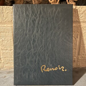 May include: A dark teal leather-bound book with the gold-coloured word "Renoir" embossed on the cover. The book has a textured surface and a rectangular shape. The background features decorative statues.