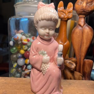 Puede incluir: Una figura de cerámica de un niño arrodillado con una túnica rosa y un lazo rosa a juego en el pelo. El niño sostiene un racimo de uvas blancas en una mano y un pequeño objeto azul en la otra. La figura tiene un acabado suave y brillante.